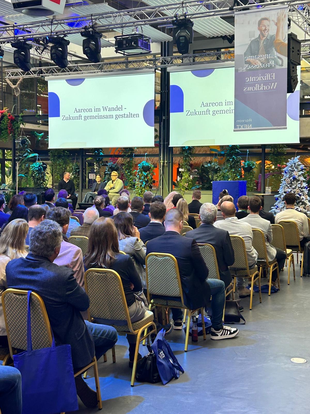 Eröffnungsmoderation mit 3 Menschen auf der Bühne und vielen Menschen im Plenum sitzend